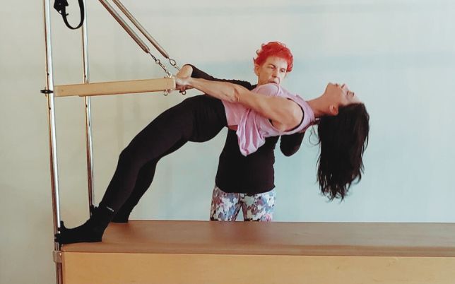 Woman balanced on Pilates Cadillac trapeze while instructor positions her shoulders.